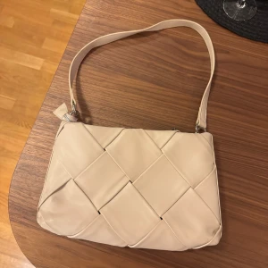 Beige axelväska med flätad design - Snygg beige axelväska med en unik flätad design. Väskan har en dragkedja upptill och en bekväm axelrem. En liten fläck på ena sidan (se bild 2) men går säkert bort efter en tvätt. 