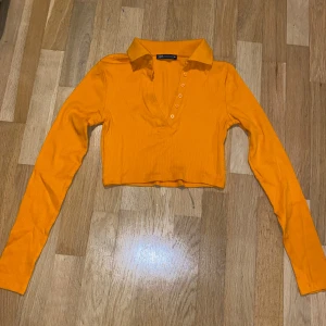 Orange ribbad croppad topp från Zara - Säljer en snygg orange ribbad topp från Zara. Den är croppad med långa ärmar och har en stilren krage med knappar framtill. Perfekt för att liva upp garderoben med lite färg!