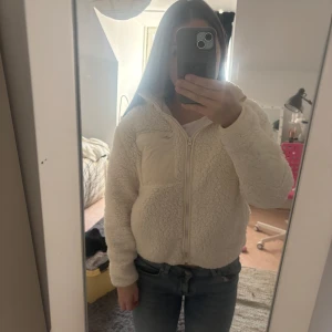 Vit teddyjacka med dragkedja - Mysig vit teddyjacka med dragkedja och en praktisk bröstficka. Perfekt för kyliga dagar när du vill hålla dig varm och bekväm. Jackan har en huva och långa ärmar.