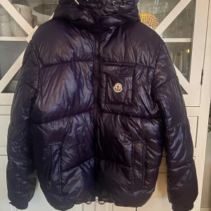 Mörkblå pufferjacka från Moncler - Säljer en snygg mörkblå pufferjacka från Moncler med huva och dragkedja. Jackan har en bröstficka med Moncler-logga och en randig detalj längs dragkedjan. Perfekt för kyliga dagar. Kontakta mig för frågor😃