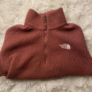 Rosa fleece från The North Face - Mysig rosa fleece från The North Face med halv dragkedja och hög krage. Perfekt för kyliga dagar. Den har en broderad logga på bröstet och är superbekväm att bära.