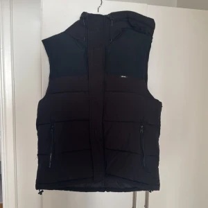 Svart dunväst från Zara - Säljer en svart dunväst från Zara med dragkedja och huva. Västen har en stilren design med två praktiska fickor framtill. Perfekt för kyligare dagar.