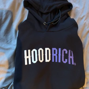 Svart hoodie från Hoodrich - Snygg svart hoodie från Hoodrich med stor logga i vitt och lila på framsidan. Perfekt för en avslappnad stil. Knappt använd då den var för stor för mig (ny skick)