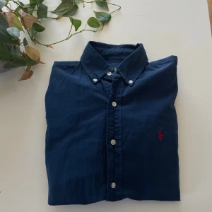 Mörkblå skjorta från Ralph Lauren - Säljer en stilren mörkblå skjorta från Ralph Lauren med klassisk krage och knappar framtill. Skjortan har en broderad logotyp i rött på bröstet. Perfekt för en elegant look. Jag skulle säga att den sitter mer som en S en än XS. Skriv vid fundering🙌