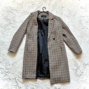 Rutig kappa  - Snygg rutig kappa i beige, svart och brun. Den har en klassisk design med långa ärmar och knappar framtill. Perfekt för höst och vår!