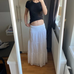 Vit långkjol med mönster - Säljer en vacker vit långkjol med subtilt mönster. Kjolen har en elastisk midja för bekväm passform och ett luftigt fall. Perfekt för varma dagar och ger en bohemisk touch till din stil. Passar bra med en enkel topp eller blus. Stl 146-152 men skulle säga att den är ganska lång och passar som en xs