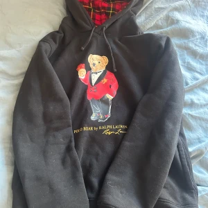Svart hoodie från Ralph Lauren - Säljer en svart hoodie från Ralph Lauren med ett coolt björnmotiv på framsidan. Hoodien har en rutig insida i huvan och dragsko. Perfekt för en avslappnad stil!