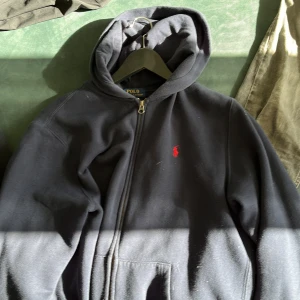 Polo Ralph Lauren Zip - Säljer en klassisk svart hoodie från Ralph Lauren med dragkedja och röd broderad logga på bröstet. Perfekt för en avslappnad stil. Huvan ger extra komfort och värme. Köpt på Kids Brand Store