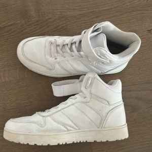 Vita sneakers med snörning och kardborreband - Snygga vita sneakers med hög modell och snörning. Skorna har även ett praktiskt kardborreband runt ankeln för extra stöd. Perfekta för en sportig look.