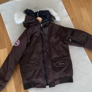 Brun dunjacka från Canada Goose - Säljer en brun dunjacka från Canada Goose med vit pälsdetalj på huvan det är en äkta Canada goose köpt i kålbåten Oslo man kan ta bort pälsen den va köpt för 10000 för några år sedan knappast använd väldigt fint skick