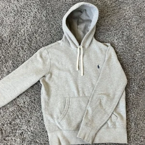 Grå hoodie från Ralph Lauren - Säljer en stilren grå hoodie från Ralph Lauren med klassisk logga på bröstet. Har ett pytte litet hål i luvan! Annars perfekt skick 