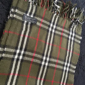 Rutig halsduk från Burberry - Snygg rutig halsduk från Burberry i grönt, svart, vitt och rött. Tillverkad i 100% ull med klassiskt mönster och fransar i ändarna. Perfekt för att hålla sig varm och stilren under kyliga dagar.