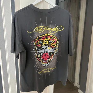 Ed Hardy T-shirt  - Säljer en grå t-shirt från Ed Hardy med ett coolt tigertryck på baksidan och gul text på framsidan. T-shirten har en oversized passform och korta ärmar. Perfekt för en casual look! Som ny i skick, endast testad och använd 1-2 gånger! Köpte för 600kr