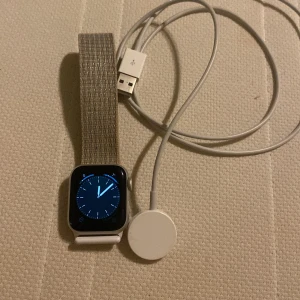 Appel Watch med grått armband - Säljer en Appel Watch  med fyrkantig skärm och grått textilarmband. Klockan har en silverfärgad boett och kommer med en laddare. Perfekt för den teknikintresserade!
