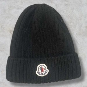 Svart Moncler Mössa - Snygg svart ribbad mössa från Moncler med deras ikoniska logotyp framtill. Perfekt för att hålla värmen under kyliga dagar. Stilren design som passar till det mesta. Hör av dig vid frågor, priset går att diskuteras!🤩