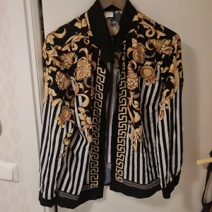 Mönstrad jacka med gulddetaljer - Säljer en unik jacka med svart och vit randigt mönster och guldfärgade barockdetaljer. Jackan har långa ärmar och en klassisk krage. Perfekt för dig som vill sticka ut med en stilren och iögonfallande look.