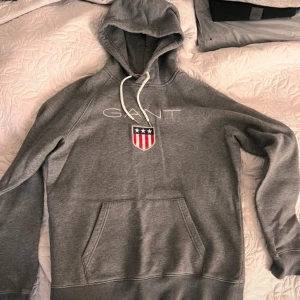Grå hoodie från GANT - Säljer en grå hoodie från GANT med en stor ficka framtill och justerbar huva med snören. Tröjan har ett broderat GANT-emblem på bröstet och är tillverkad i mjukt material för extra komfort.