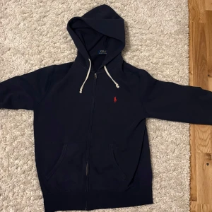 Svart hoodie från Polo Ralph Lauren - Säljer en stilren svart hoodie från Polo Ralph Lauren med dragkedja och en liten röd logga på bröstet. Perfekt för en avslappnad look. Den har en justerbar huva och praktiska fickor framtill. Passar både för vardag och chill. Priset kan diskuteras 