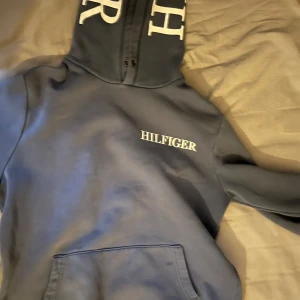 Blå hoodie från Tommy Hilfiger - Säljer en snygg blå hoodie från Tommy Hilfiger med stor logga på bröstet och huvan. Perfekt för en avslappnad stil. Hoodien har en klassisk känguruficka och justerbar huva med snören.
