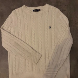 Vit stickad tröja från Polo Ralph Lauren - Säljer en stilren vit stickad tröja från Polo Ralph Lauren med klassiskt flätmönster. Tröjan har långa ärmar och en broderad logotyp på bröstet. Perfekt för en elegant och avslappnad look.