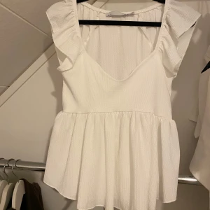 Vit blus med volangdetaljer - Säljer en söt vit blus med volangdetaljer på axlarna och en v-ringad design. Blusen har en luftig passform och är perfekt för varmare dagar. Den är i ett lätt och bekvämt material.