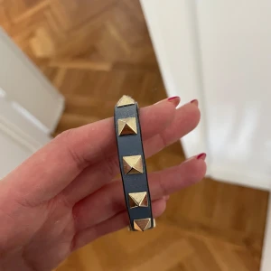 Blått armband med nitar från Valentino - Snyggt svart armband från Valentino dekorerat med guldiga pyramidformade nitar. Perfekt accessoar för att ge en edgy touch till din outfit. Kommer i en elegant röd förpackning. Nypris runt 2500kr