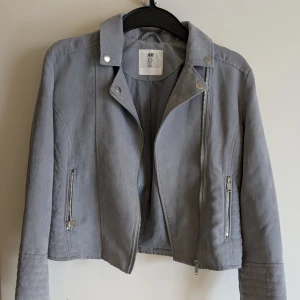 Grå jacka från H&M - Snygg grå jacka från H&M med dragkedja och stilrena detaljer. Jackan har en klassisk bikerstil med långa ärmar och silverfärgade knappar. Perfekt för en cool och avslappnad look. 100% Polyester. Tvättas i 40 grader. Min dotters namn är tillfälligt över klistrat med ett vitt klistermärke. Jag kommer att stryka över hennes namn med svart textil penna när jackan säljs.