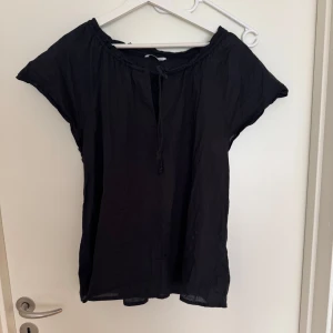 Svart blus från Ellos - Säljer en stilren svart blus från Ellos med korta ärmar och knytband vid halsen. Perfekt för en avslappnad look. Blusen är tillverkad i ett lätt och luftigt material.