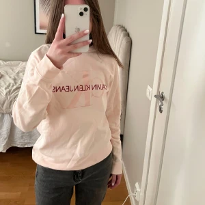Ljusrosa sweatshirt från Calvin Klein - Säljer en stilren ljusrosa sweatshirt från Calvin Klein Jeans med deras ikoniska logga tryckt på framsidan. Tröjan har långa ärmar och en rund halsringning. Perfekt för en avslappnad look.