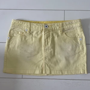 Gul kortkjol i jeansmaterial - Säljer en söt gul kortkjol i jeansmaterial med klassisk femficksdesign. Kjolen har en knapp och dragkedja framtill samt bälteshällor. Perfekt för en solig dag!