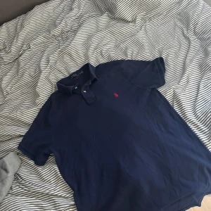 Mörkblå pikétröja från Ralph Lauren - Säljer en klassisk mörkblå pikétröja från Ralph Lauren med den ikoniska röda loggan på bröstet. Tröjan har korta ärmar och en stilren krage med knappar. Perfekt för en avslappnad men ändå stilren look.