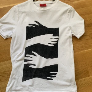 Vit t-shirt från Hugo Boss - Säljer en stilren vit t-shirt från Hugo Boss med ett unikt svart tryck av händer på framsidan. T-shirten är kortärmad och tillverkad i mjuk bomull. Perfekt för en casual look. Unisex-modell.
