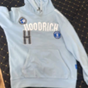 Ljusblå hoodie från Hoodrich - Säljer en ljusblå hoodie från Hoodrich med stora vita bokstäver på bröstet och coola blå patches. Tröjan har huva med snörning, känguruficka och ribbade muddar. Perfekt för dig som gillar streetwear och vill sticka ut med snygga detaljer.