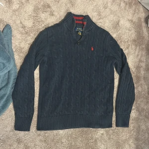 Mörkblå stickad tröja Polo Ralph Lauren - Mörkblå kabelstickad tröja från Polo Ralph Lauren med halv dragkedja och ståkrage. Insidan av kragen har röda ränder och tröjan har en röd broderad logga på bröstet. Perfekt för dig som gillar klassisk och stilren look. Passar dig som är 164-175 