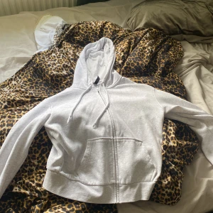 Grå hoodie med dragkedja från H&M - Ljusgrå hoodie från H&M Divided i storlek M. Tröjan har dragkedja framtill, snörning i huvan och en stor magficka. Tillverkad i mjuk bomullsblandning och har en klassisk, avslappnad passform med långa ärmar.