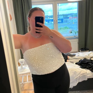 Vit glittrig bandeau tubtopp - Säljer en vit bandeau tubtopp täckt med glittriga paljetter. Toppen är axelbandslös och croppad, perfekt för dig som vill sticka ut. Snygg att matcha med både jeans och kjol för en cool look.