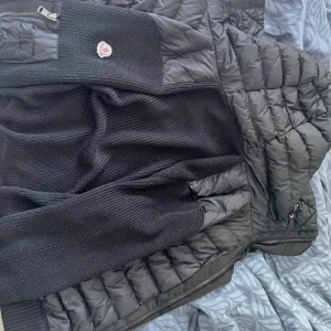 Svart Moncler pufferjacka med stickade ärmar - Säljer en svart pufferjacka från Moncler med stickade ärmar och detaljer. Jackan har dragkedja framtill, huva och en ficka med dragkedja på ärmen. Klassisk Moncler-logga på ärmen och quiltad kropp för extra värme. Perfekt för kalla dagar.