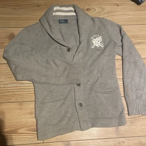 Grå kofta från Polo Ralph Lauren - Säljer en grå stickad cardigan från Polo Ralph Lauren med broderad logga på bröstet. Den är gjord av ett riktigt skönt tyg och har sjukt bra passform. Köptes för ca 4500kr och säljes för endast 650kr:)