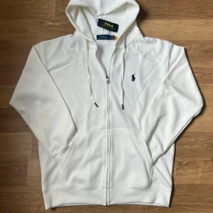 Vit hoodie från Polo Ralph Lauren - Snygg vit hoodie från Polo Ralph Lauren med klassisk broderad logga på bröstet. Tröjan har huva med snörning, dragkedja framtill och två fickor. Perfekt för en clean och stilren look. Tillverkad i mjuk bomull för maximal komfort.