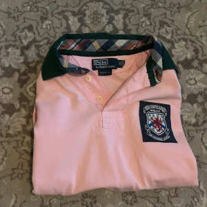 Rosa pikétröja Polo Ralph Lauren - Snygg rosa pikétröja från Polo Ralph Lauren med grönt krage och rutigt tyg på insidan. Broderad patch på bröstet och stort vitt nummer 14 på ryggen. Klassisk passform och korta ärmar, perfekt för en sportig och preppy stil.
