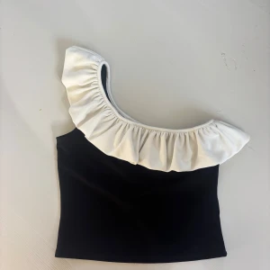 Svart och vit oneshoulder topp med volang - Snygg svart topp med bred vit volang längs en axel. Oneshoulder-modell som ger en cool och trendig look. Perfekt för dig som vill sticka ut med en unik och stilren topp. Mjuk och skön att bära.