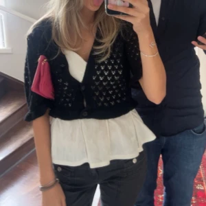 Svart kort stickad kofta med knappar - Säljer en svart, kort och stickad kofta med knappar framtill. Koftan har ett hålmönster och är perfekt att slänga över en topp. Den har korta ärmar och en loose passform som ger en avslappnad vibe.