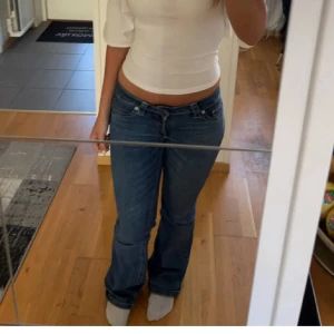 Blå bootcut jeans med mönstrade fickor - Säljer ett par blå bootcut jeans med låg midja och coola vita mönster på bakfickorna. Jeansen har klassisk femficksdesign och är tillverkade i jeansmaterial. Perfekta för dig som vill sticka ut lite extra med detaljer på baksidan.