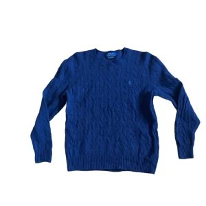 Ralph Lauren kabelstickad tröja  - Kabelstickad blå tröja från Polo Ralph Lauren i klassisk modell med rund hals och diskret logga på bröstet. Tröjan är långärmad och gjord i mjuk bomull, perfekt för lager-på-lager under höst och vinter. Snygg ribbad kant vid ärmslut och nederkant. Har 2 minde hål som syns på de sista 2 bilderna (lätt att sy om man kan det) skriv för att få bild på måtten !