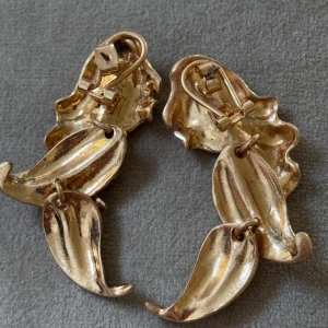 Guldiga statement örhängen i bladform - Unika örhängen i guldtonad metall med coola, organiska bladformer och små glittriga stenar längs ena kanten. De har en snygg vintagevibe och är perfekta för att sticka ut. Klämfäste för enkel användning.