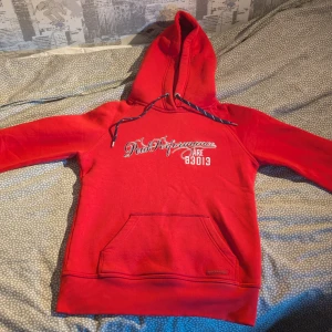 Röd Peak Performance hoodie med tryck - Snygg röd hoodie från Peak Performance med vit-svart broderad logga och text på bröstet. Tröjan har huva med snörning, magficka och ribbade muddar. Tillverkad i mjuk bomulls- och polyestermix, perfekt för chill eller utomhusaktiviteter.