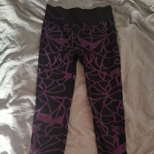 Svarta och lila leggings från Panos - Snygga svarta leggings från Panos med lila abstrakt mönster. De är tillverkade i snabbtorkande syntetmaterial och har hög midja för extra komfort. Perfekta för träning eller chill dagar. Tight passform och elastisk midja. Hela byxorna är väldigt elastiska och sitter bekvämt runt benen :)