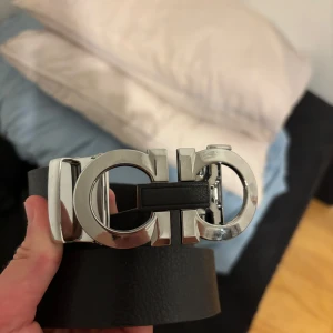 Svart Ferregamo liknande skärp med silverdetalj - Säljer ett svart skärp från ett Ferragamo liknande företag med en bred rem i skinn och en ikonisk dubbel Gancini-spänne i blankt silver. Skärpet har en stilren och lyxig känsla, perfekt för att lyfta vilken outfit som helst.