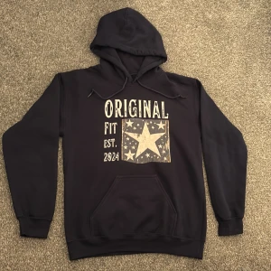Mörkblå hoodie med stjärntryck - Mörkblå hoodie med stor vit stjärna och texten 'ORIGINAL FIT EST. 2024' på bröstet. Klassisk känguruficka, huva med snörning och lång ärm. Materialet är en bomullsblandning som känns mjuk och skön. Perfekt för dig som gillar hoodies med tryck. Säljer den då den är för liten för mig. Passar bäst för 165cm-175cm killar. Tveka inte att ställa frågor!