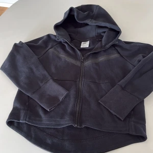 Nike tech 137/146 - Svart Nike tech-hoodie med hel dragkedja framtill. Tröjan har långa ärmar, två fickor och en diskret randdetalj över bröstet. Tillverkad i mjuk bomullsmix, perfekt för chill eller träning. Klassisk och enkel design som funkar året runt. Storlek M, 137-146